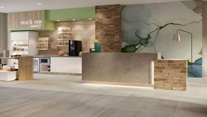 A modern reception area with a wooden reception desk, a green wall with a 'nice & vice MARKET' sign, and a large abstract mural. - Wyndham Garden Tampa Fairgrounds/Casino (Tampa)