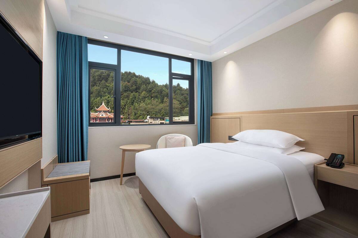 Une chambre d’hôtel avec un grand lit, une table de chevet avec un téléphone, un petit bureau avec une chaise, un téléviseur sur le côté gauche et une fenêtre donnant sur un immeuble et des arbres.