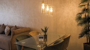 A modern living room with a glass-top dining table, green chairs, a sofa with patterned cushions, a hanging light fixture, and a potted plant.