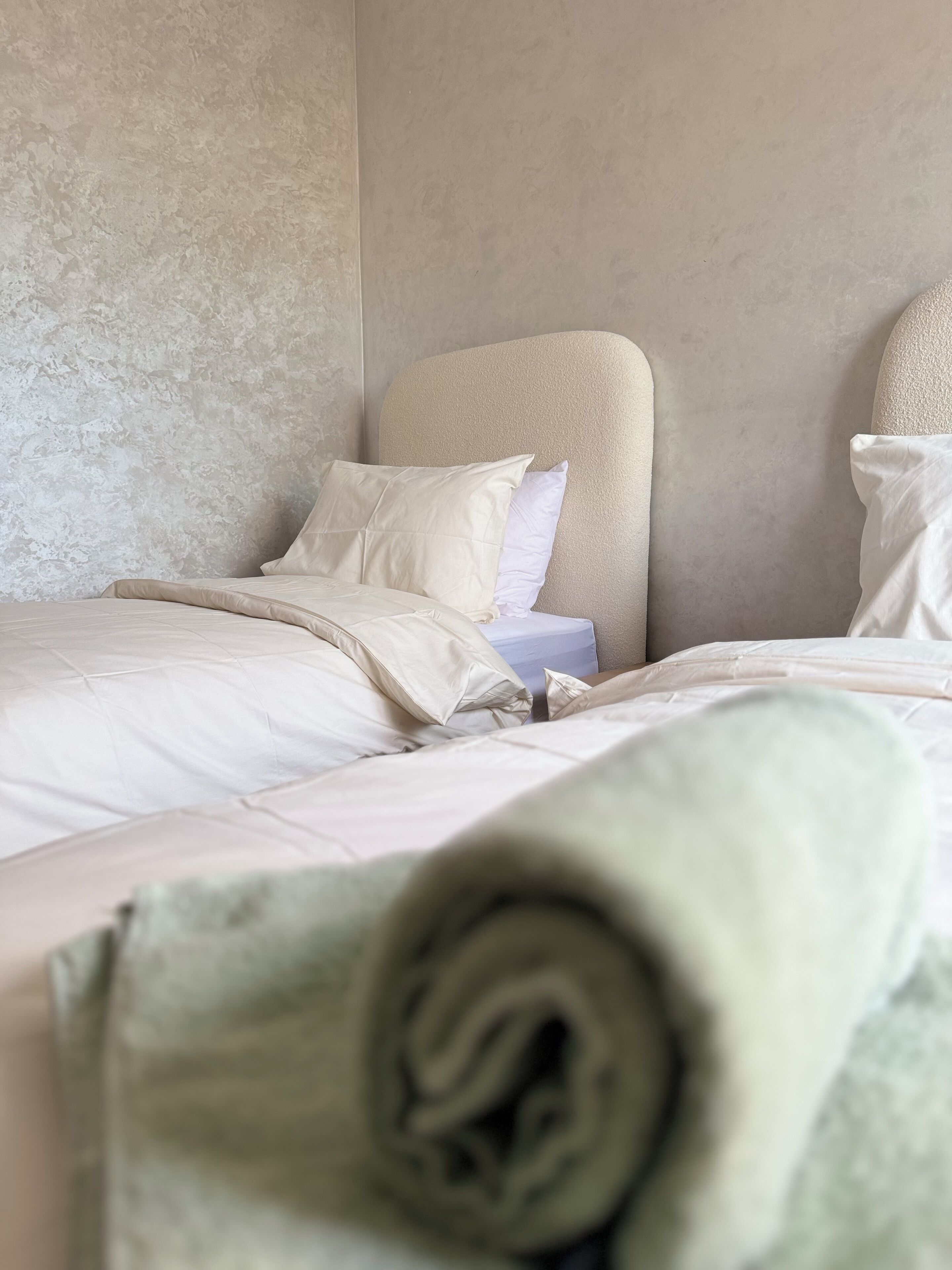 A neatly made bed with a rolled-up towel in the foreground, a headboard, and a textured wall.