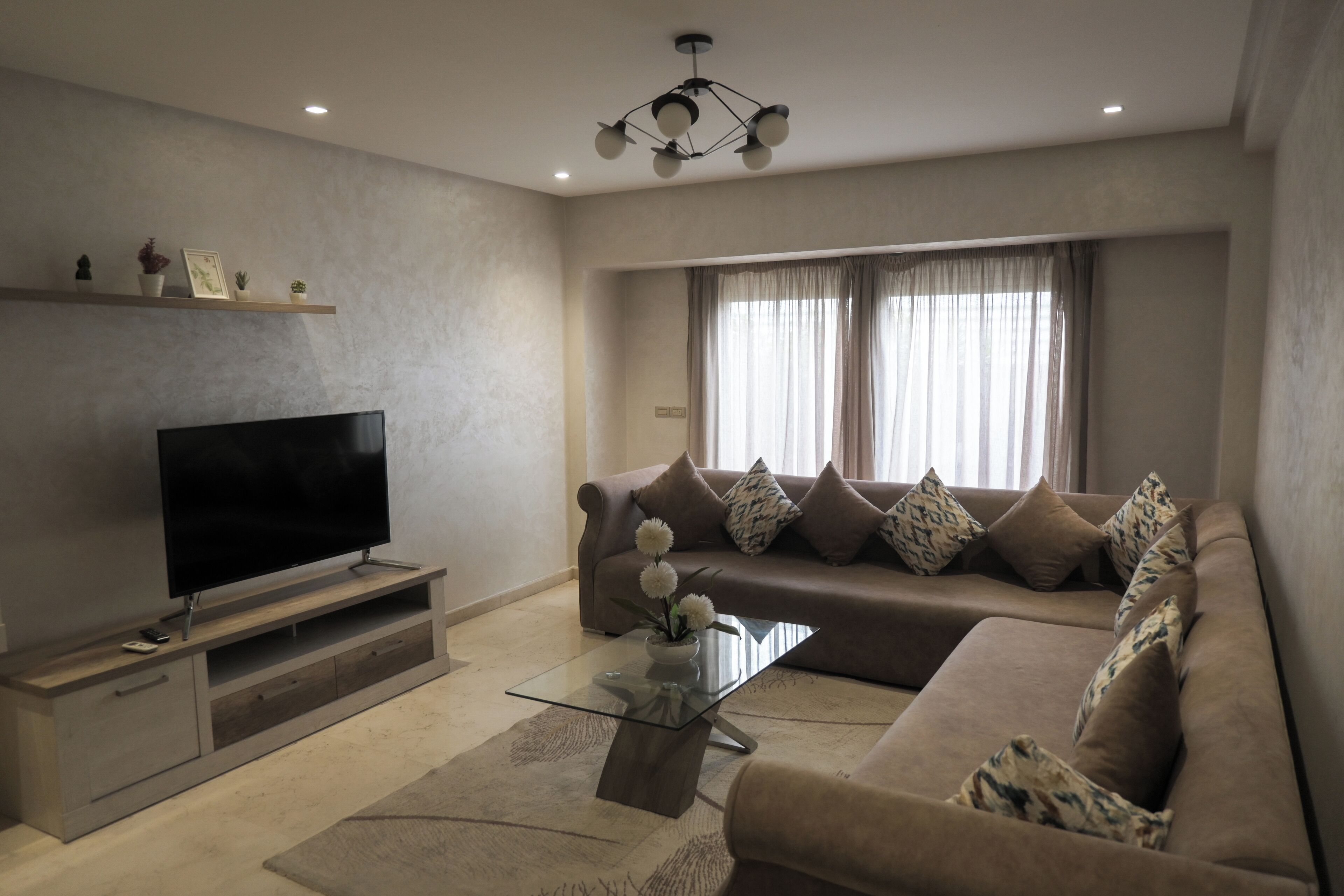 A modern living room with a flat-screen TV, a glass coffee table, a beige sofa, and a large window with curtains.