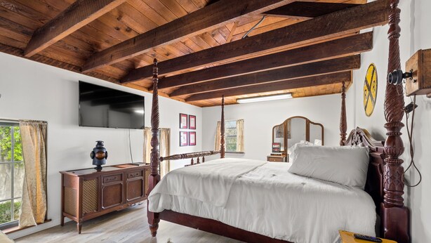 Ein Schlafzimmer mit Himmelbett, Holzbalkendecke, Flachbildfernseher, Holzkommode und einem großen Fenster mit Vorhängen.