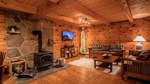 A cozy cabin interior with a stone wall, wooden furniture, a fireplace, and a Christmas tree.