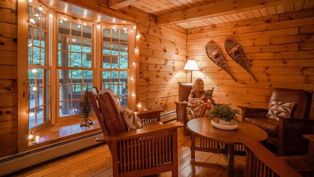 A cozy cabin interior with wooden walls, a large window with lights, a table, and two chairs.