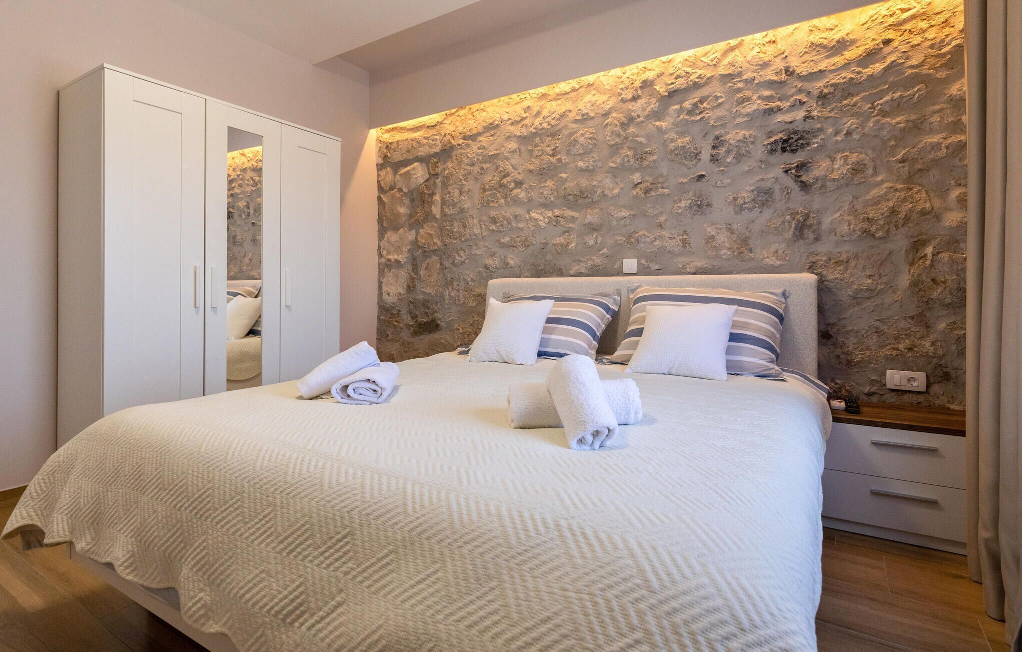 Un dormitorio con pared de piedra, una cama con almohadillas a rayas y un armario blanco.