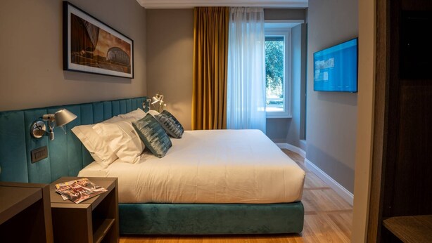 A bedroom with a bed, a nightstand, a lamp, a painting, a window with curtains, and a TV. - Your room like in a 4-star Hotel (Roma)