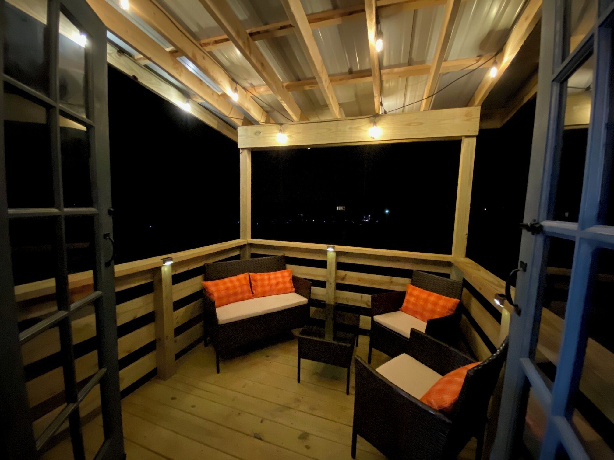 Een overdekt terras met houten meubels en kussens, uitzicht op de nachthemel en een hekwerk.