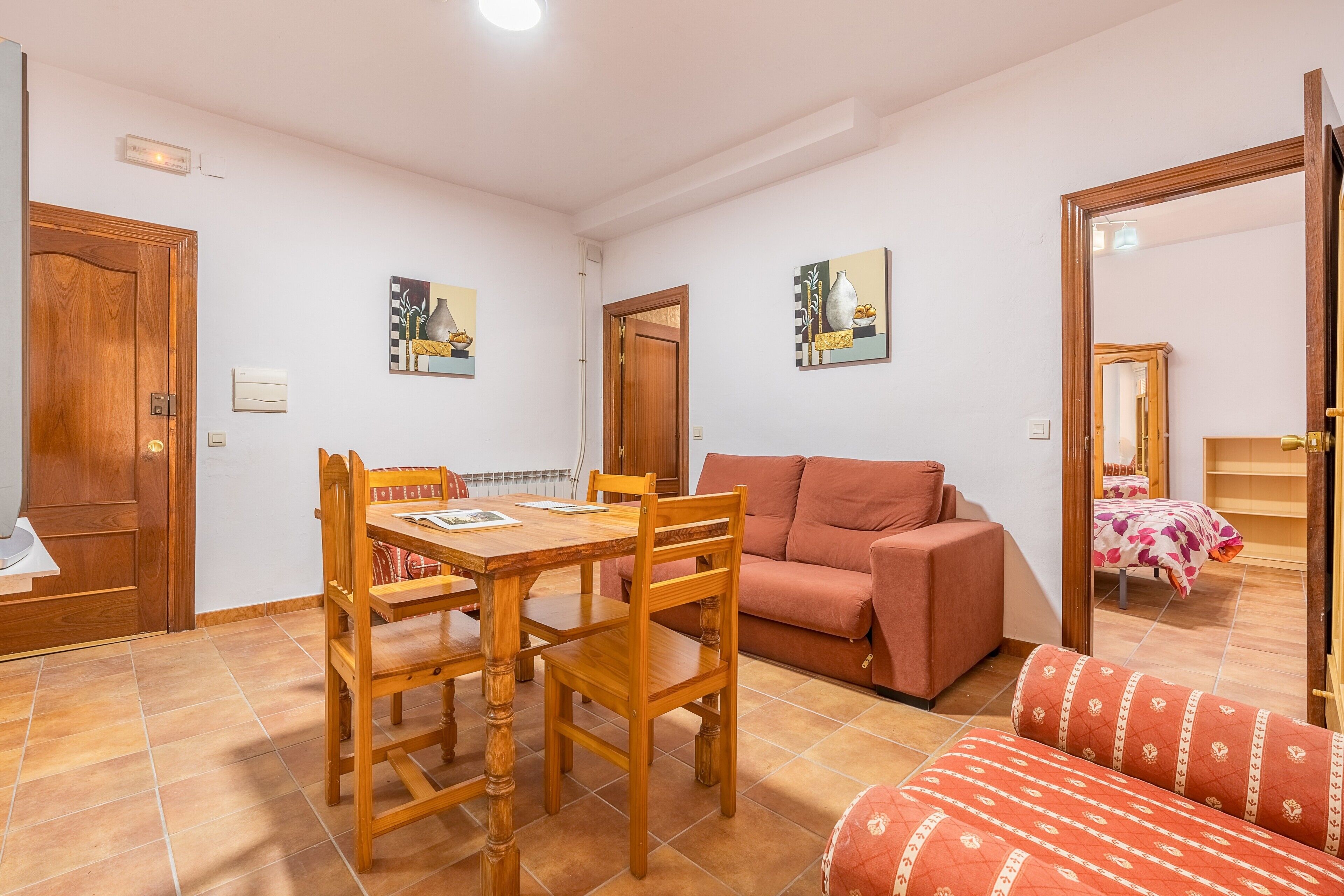 A room with a wooden dining table, a sofa, a patterned armchair, and two paintings on the walls.