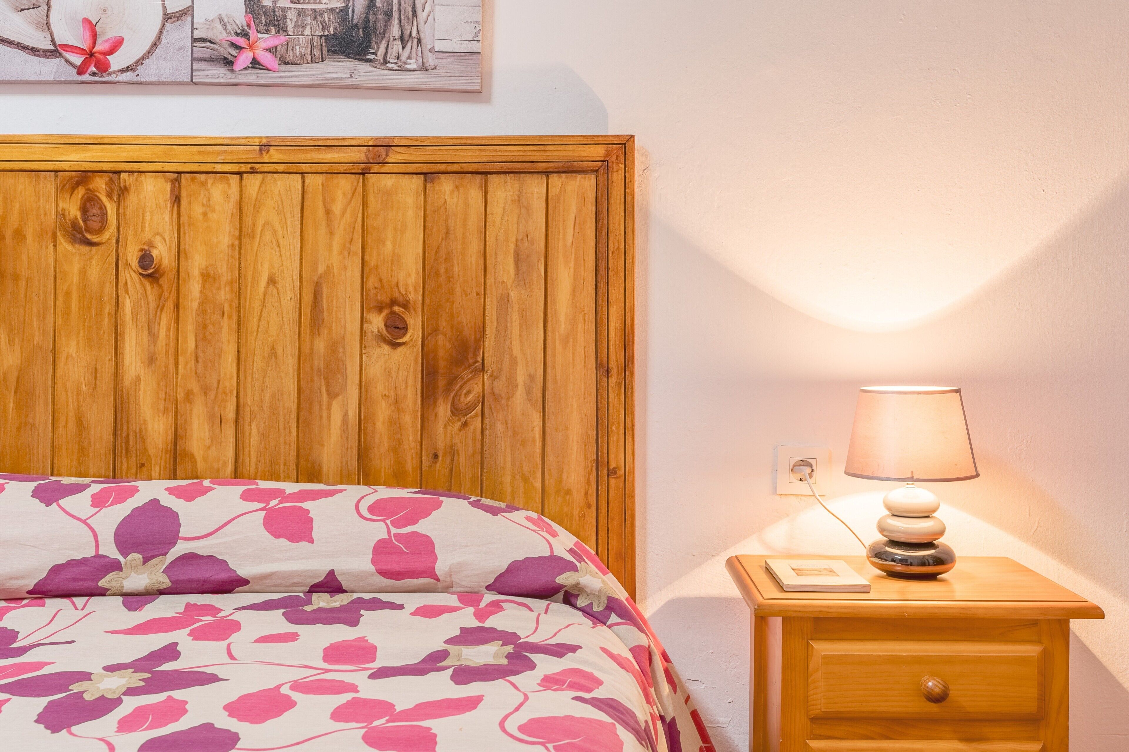 A wooden headboard, a bed with floral bedding, a bedside lamp, and a nightstand with a book.
