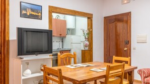 Un coin repas avec une table en bois, des chaises, un téléviseur et une cuisine en arrière-plan.