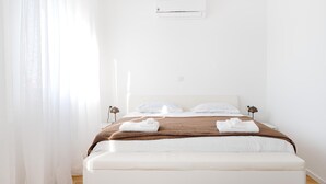 Ein helles, minimalistisches Hotelzimmer mit Bett, weißen Vorhängen und einer Deckenklimaanlage.