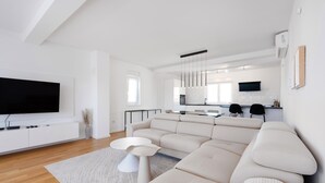 Ein modernes Wohnzimmer mit einem großen Ecksofa, einem Flachbildfernseher und einer minimalistischen Küche im Hintergrund.