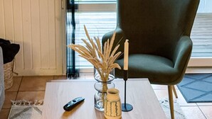 Ein Zimmer mit einem dunkelgrünen Sessel, einem kleinen Tisch mit einer Vase getrockneter Pflanzen, einer Fernbedienung und einem gemusterten Teppich.