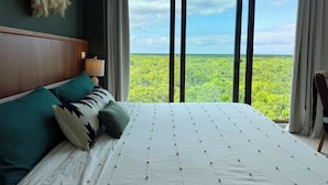 A bedroom with a large bed, a nightstand, a lamp, and a view of a forest through floor-to-ceiling windows. - Bosques de Ciudad Mayakoba (Playa del Carmen)