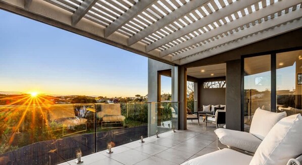 A modern outdoor balcony with a glass railing, a sofa, and a view of the sunset.