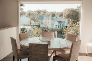 A round table with wicker chairs and a metal bowl centerpiece, set against a window overlooking a residential area with houses and trees.