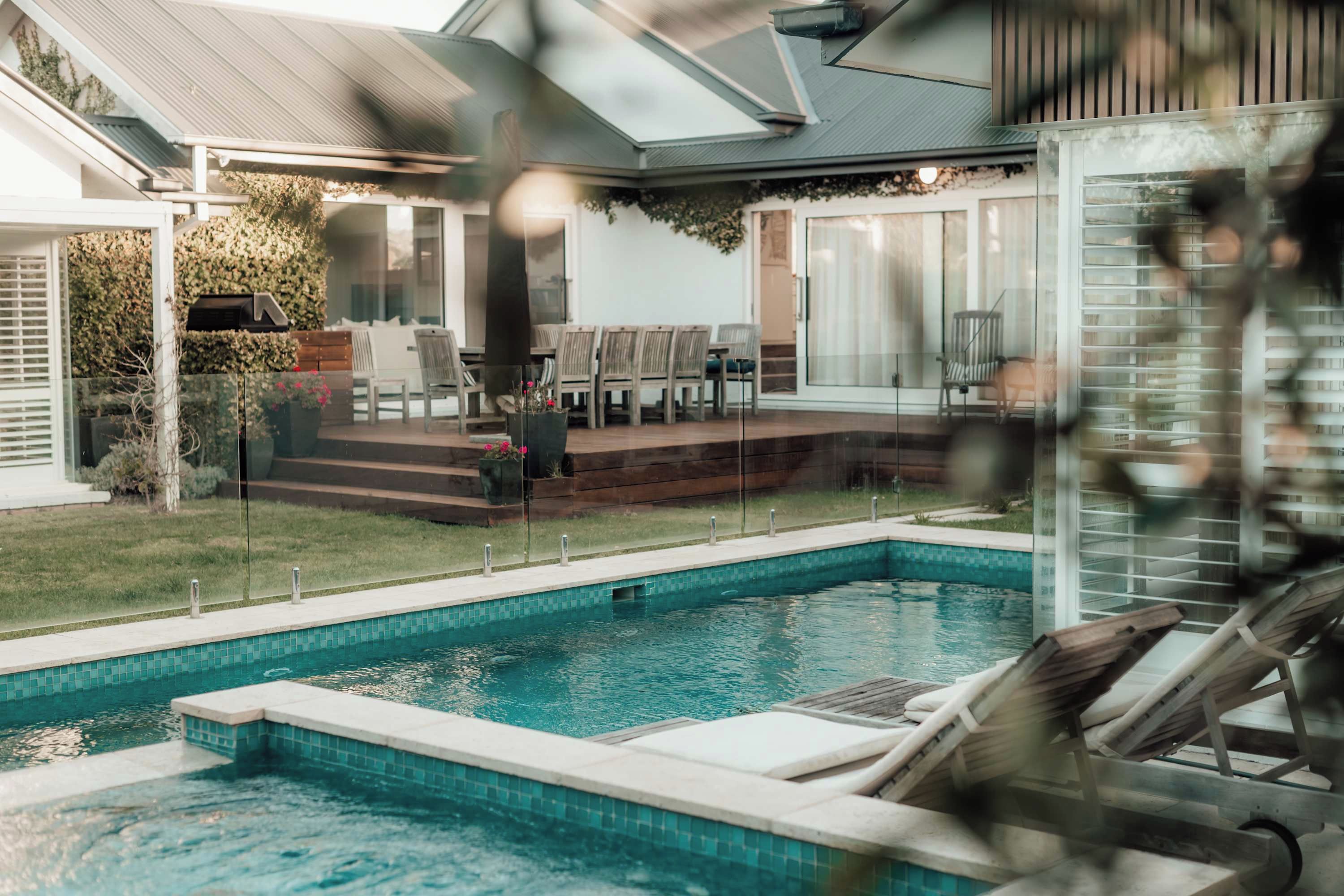 Una piscina amb zona de solàrium, envoltada per una casa amb terrassa coberta i vista al jardí de l'habitatge.