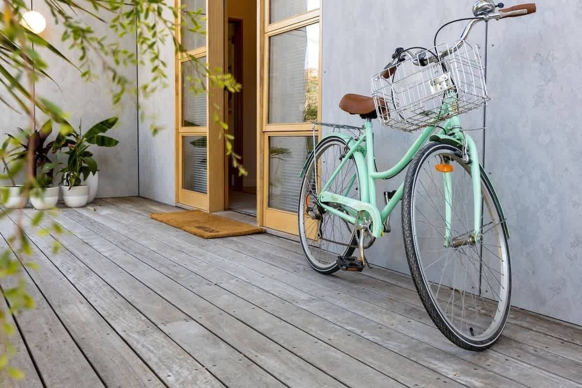 Uma bicicleta azul-turquesa com cesta, um alpendre de madeira e plantas em vasos.