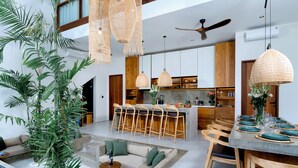 Uma sala de estar ampla com teto alto, cozinha com balcão e sala de jantar com mesa posta para quatro pessoas.