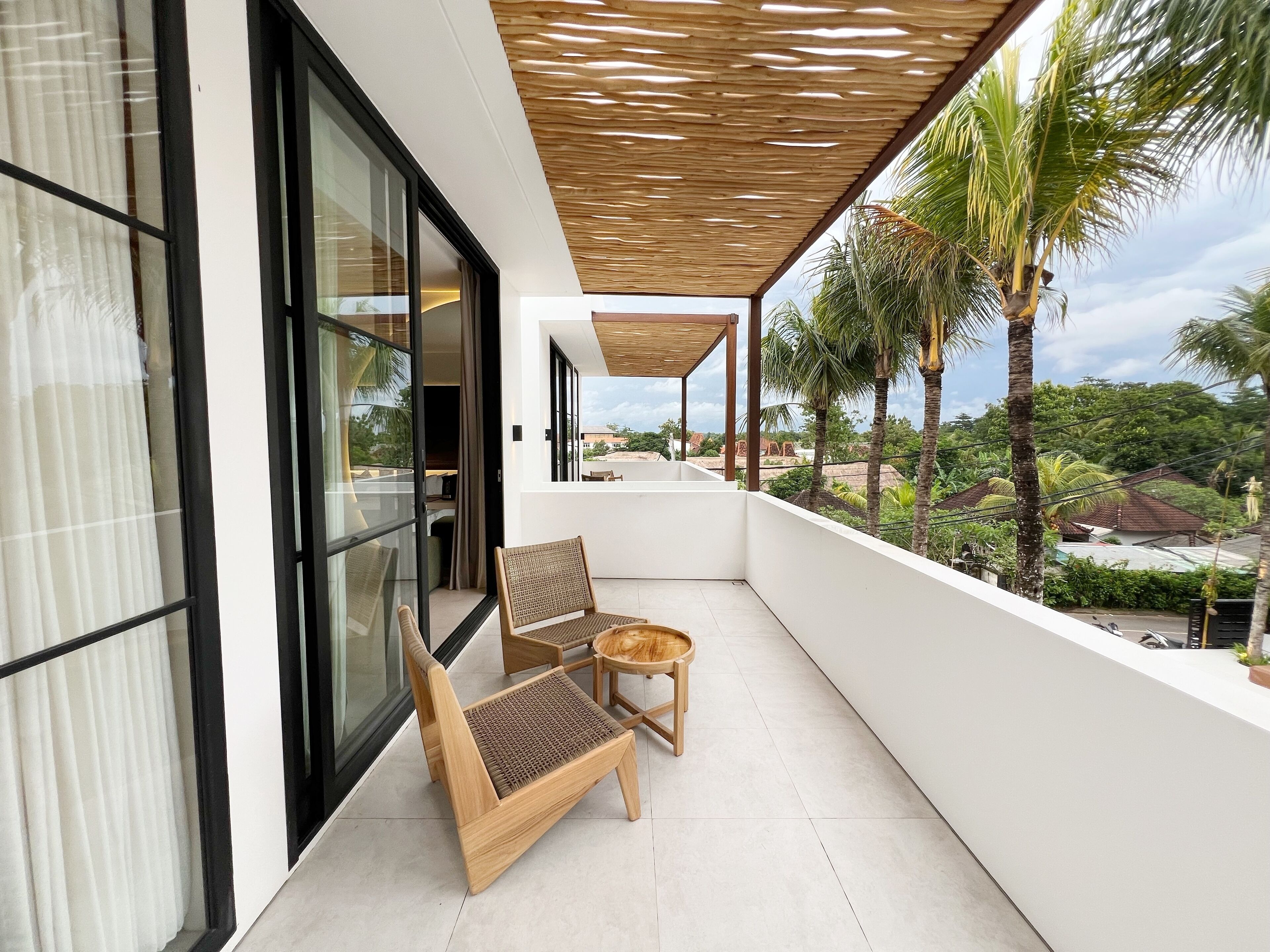 A balcony with two wicker chairs and a small table, overlooking a lush landscape with palm trees and a distant view of buildings.