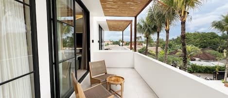 A balcony with two wicker chairs and a small table, overlooking a lush landscape with palm trees and a distant view of buildings.