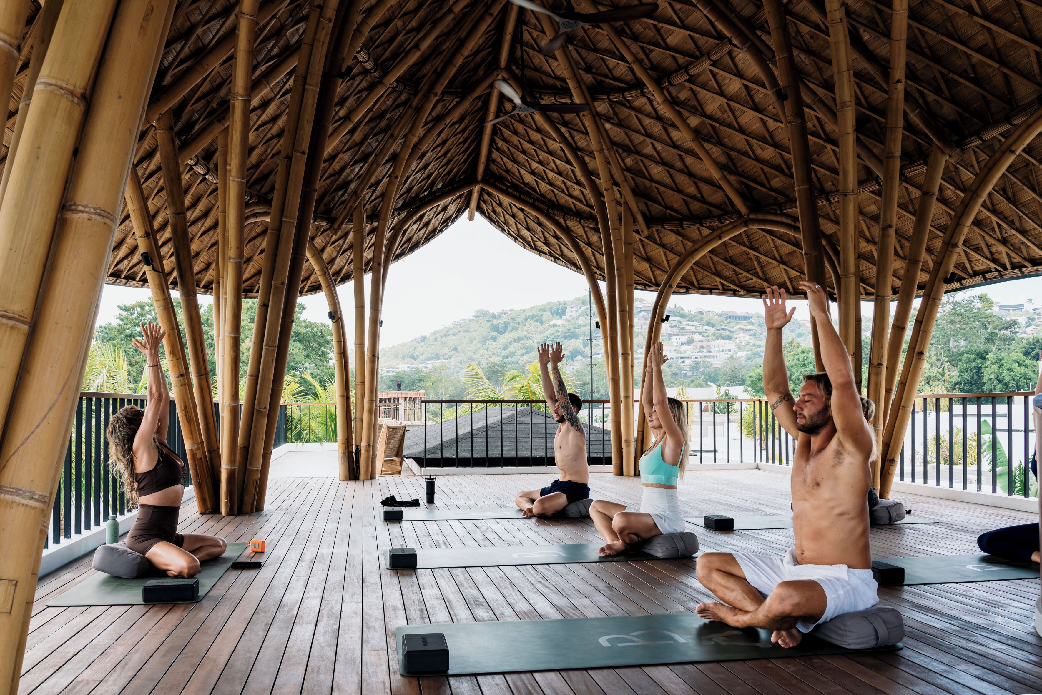 A group of people practicing yoga on a wooden deck with a bamboo roof structure.