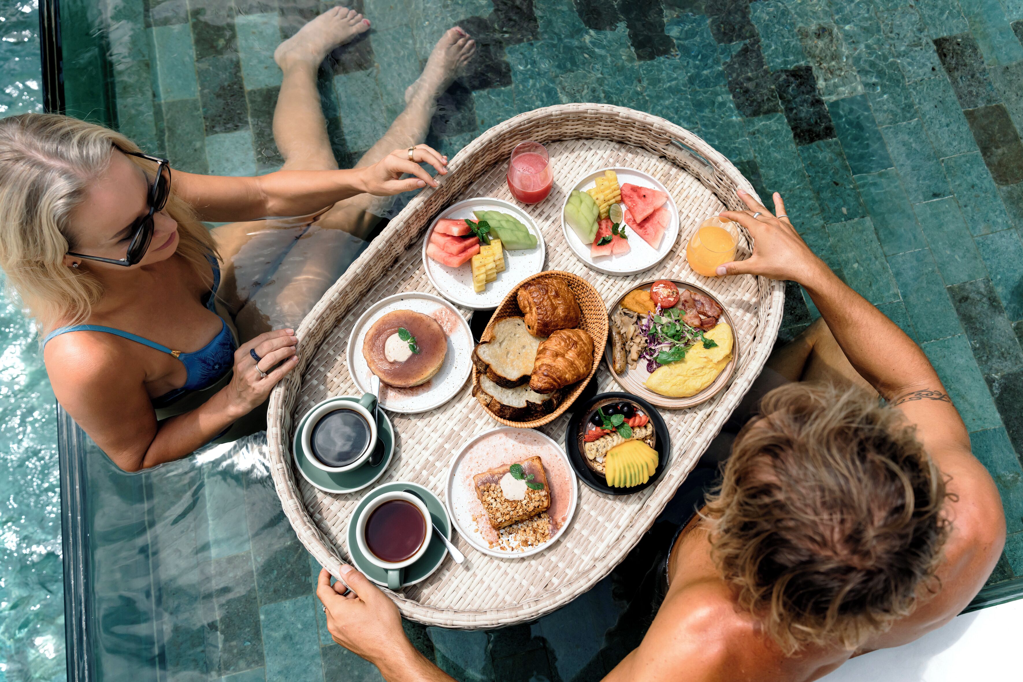 Una mesa de comedor con diversos alimentos y bebidas, entre ellos, rebanadas de sandía, mazorcas de maíz y un croissant, además de tazas de café y jugo.