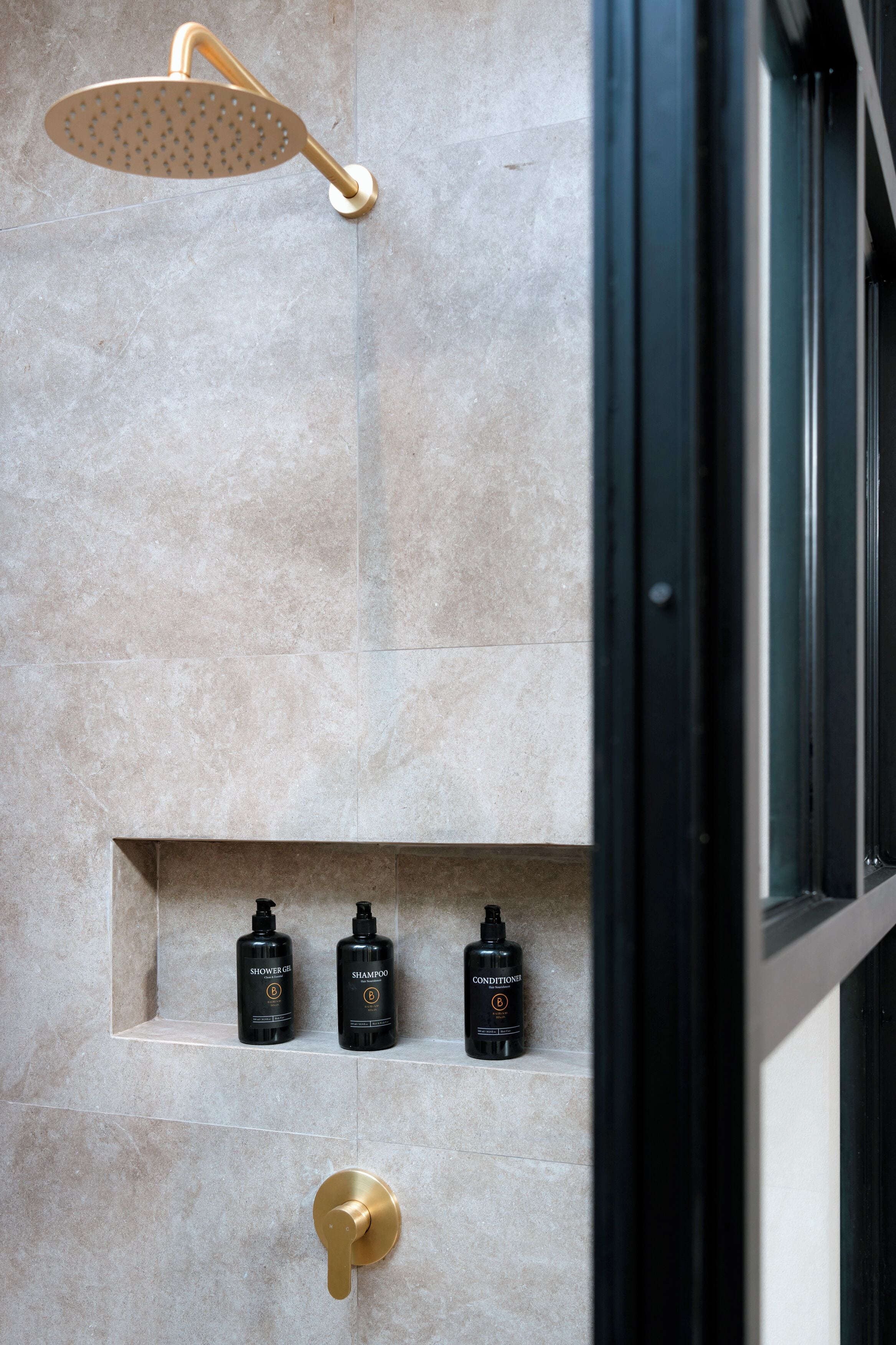 A modern shower with a gold showerhead and a gold faucet, mounted on a tiled wall. There are three black bottles of shower products on a shelf below.