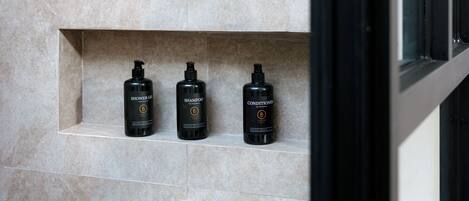 A modern shower with a gold showerhead and a gold faucet, mounted on a tiled wall. There are three black bottles of shower products on a shelf below.