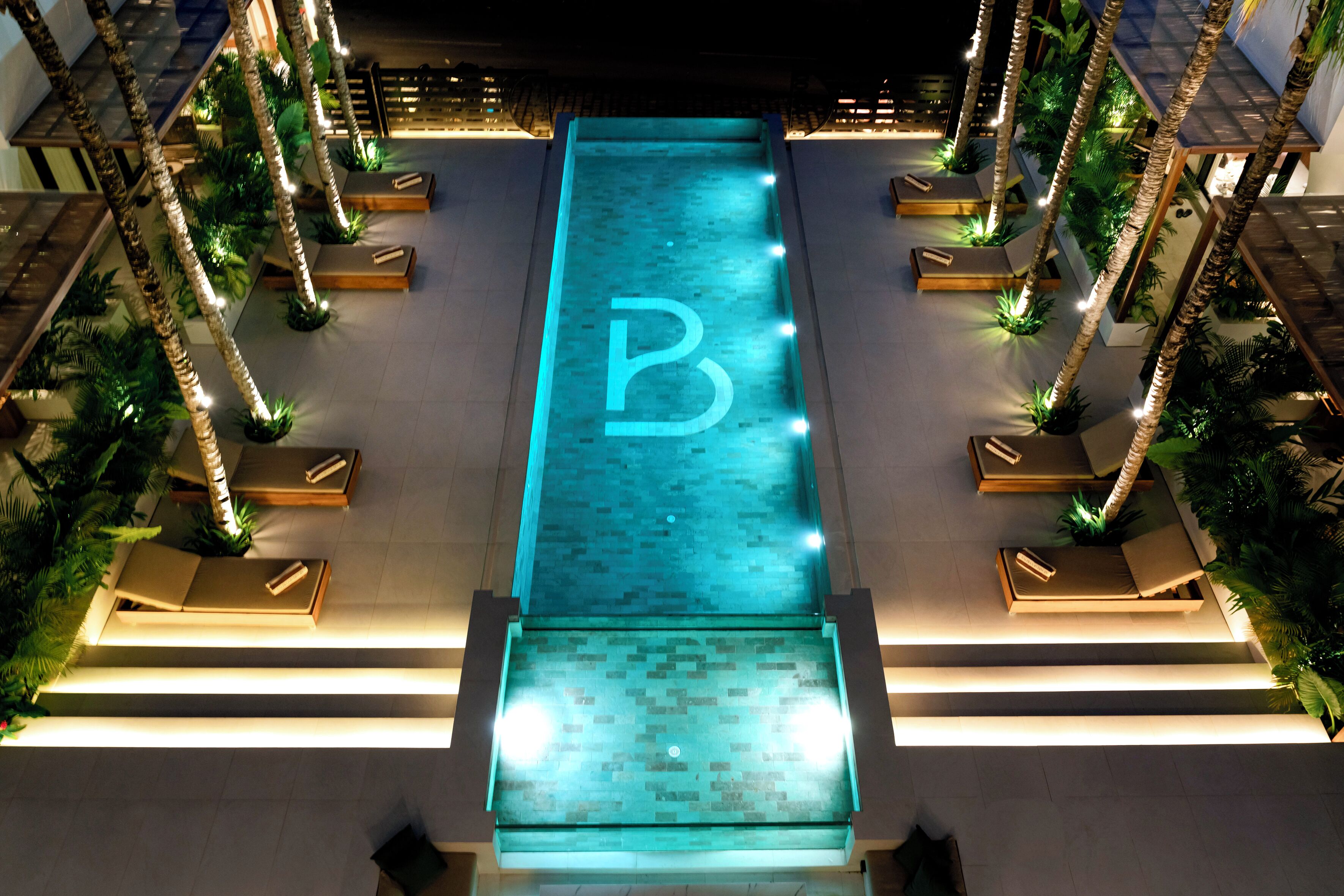 Una piscina amb un gran logotip de la lletra 'B', envoltada de palmeres i gandules.