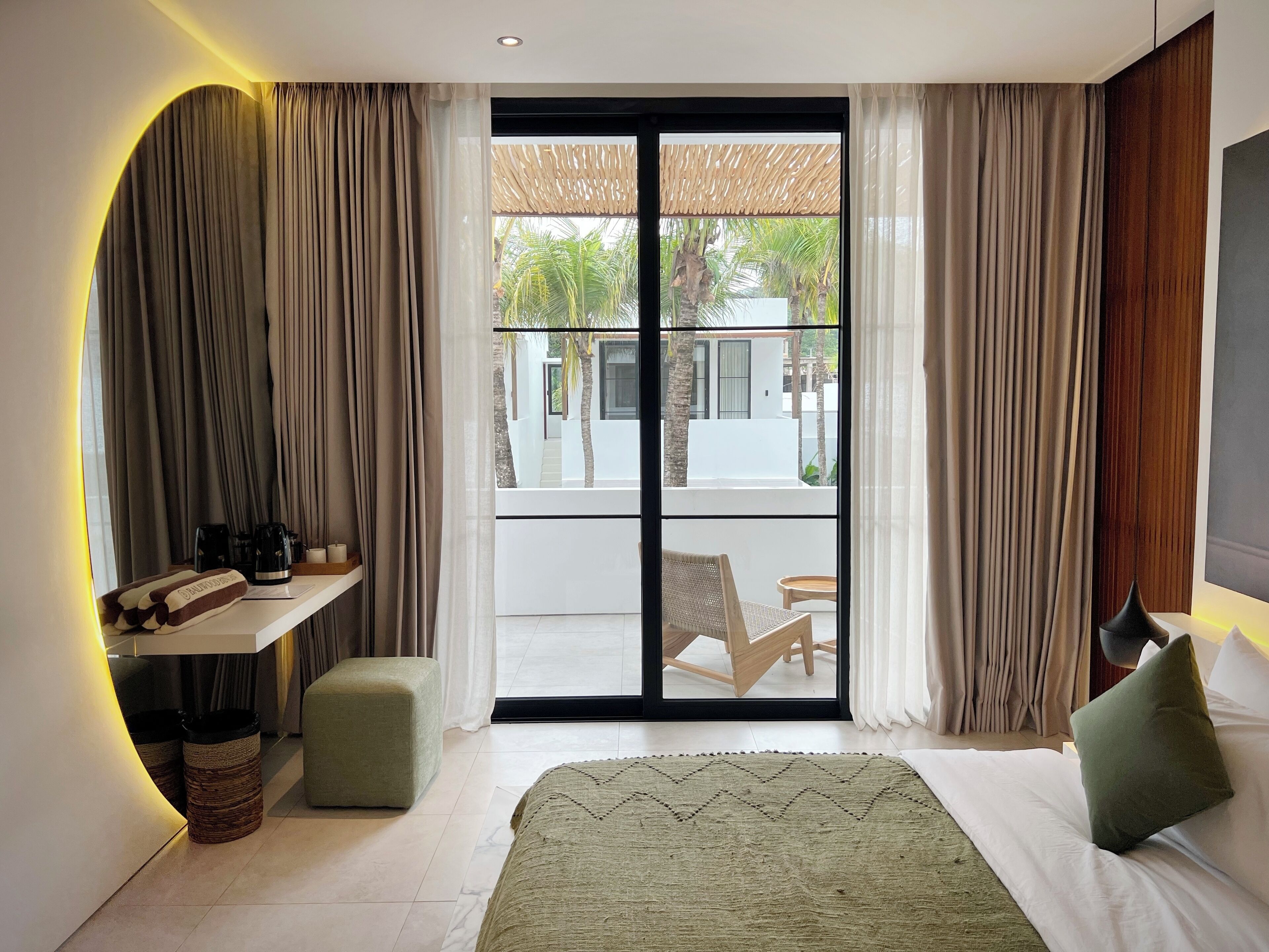 A modern bedroom with a large bed, a small seating area, and a view of a courtyard with palm trees.