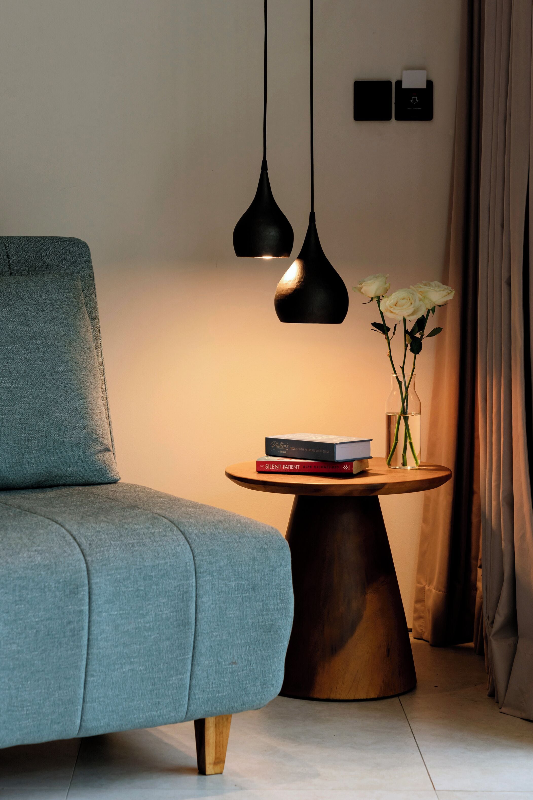 A modern living room with a grey sofa, a wooden coffee table with a vase of white flowers, and two black pendant lights.
