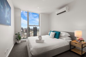 Ein modernes Hotelzimmer mit einem großen Fenster und Blick auf die Stadt, einem ordentlich bezogenen Bett mit weißen und türkisen Kissen sowie einem Nachttisch mit Lampe und Blumen.