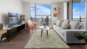 A modern living room with a large TV, a sofa, a chair, and a view of a cityscape through large windows. - 888 Collins (Docklands)