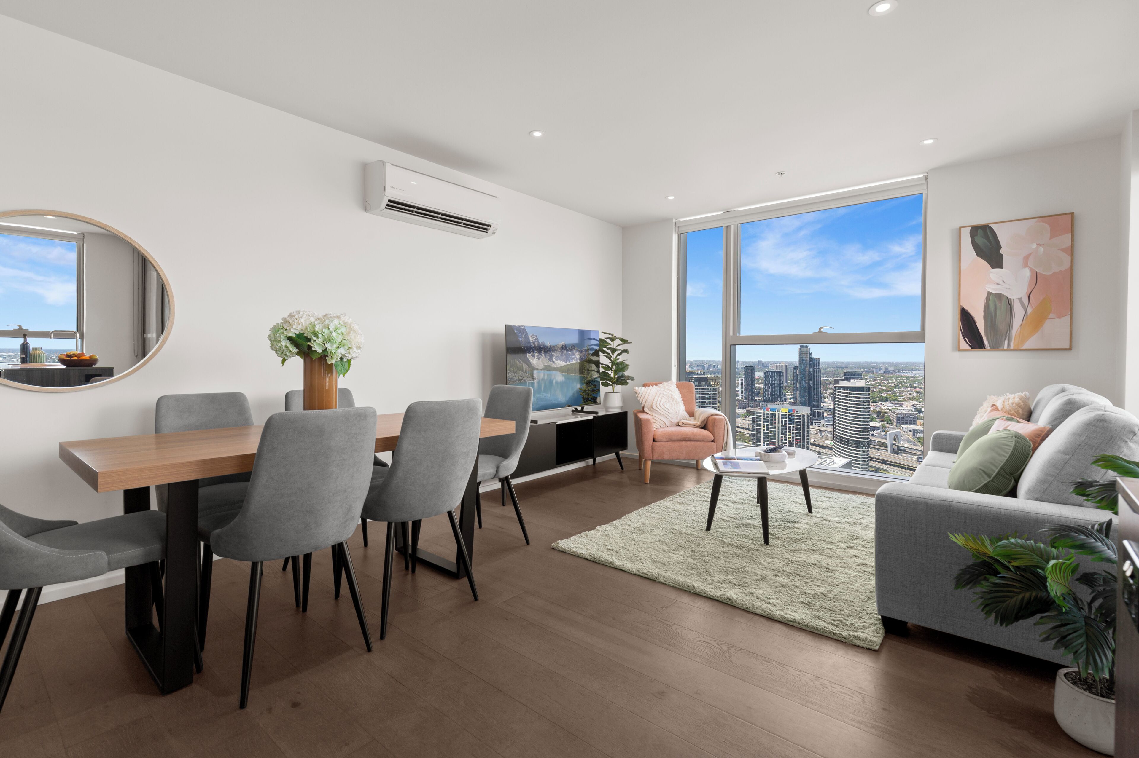 A modern living room with a dining area, large windows offering a city view, and a flat-screen TV.