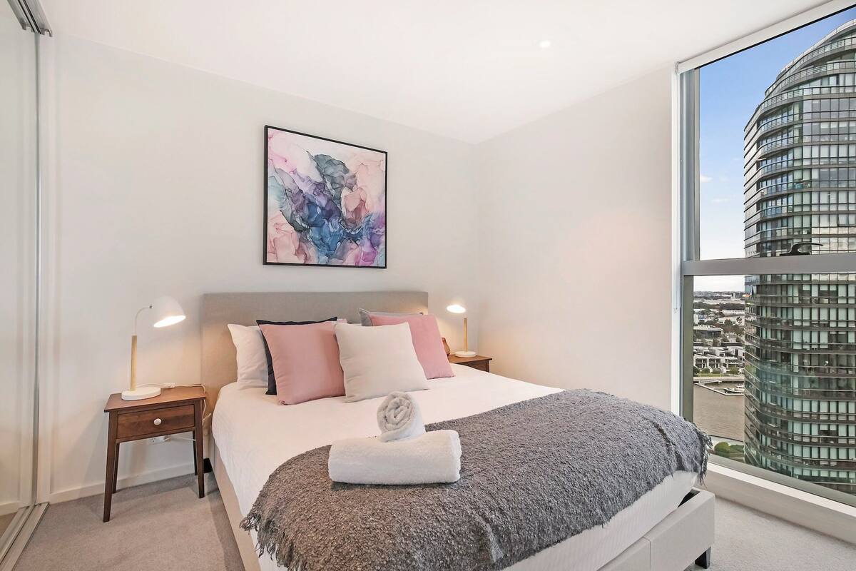 A modern bedroom with a large bed, a city view, and a painting on the wall.
