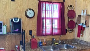 A compact kitchen with wooden walls, a clock, a coffee maker, a sink, and a window with curtains.