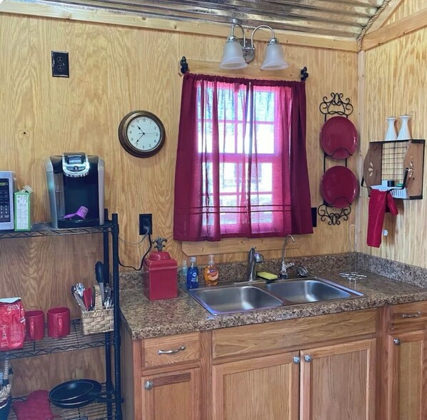 Una cocina compacta con paredes de madera, un reloj, una cafetera, un lavamanos y una ventana con cortinas.
