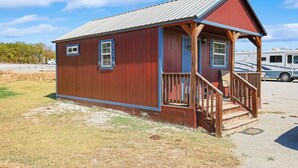 A small wooden cabin with a porch and steps, located in a rural area with a clear sky and a parked RV in the background.