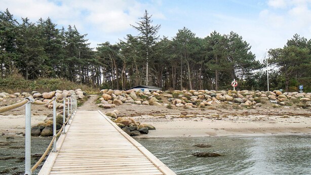 A wooden pier extending into calm water, surrounded by a rocky shoreline and dense forest. - Holiday Home With Nature View (Gørlev)