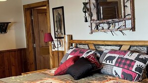 Una cama con colcha, almohadas, cabecera de madera, espejo en la pared y ventilador de techo.