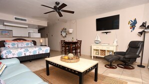 A living room with a sofa, a coffee table with a basket, a dining table with chairs, a TV, a ceiling fan, and a TV stand with a lamp. - Casa Playa Resort #102 King Suite Beach Front - "Starfish" (Fort Myers Beach)