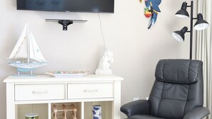 A modern living room with a flat-screen TV, a white console table with decorative items, a black leather chair, and a floor lamp. - Casa Playa Resort #102 King Suite Beach Front - "Starfish" (Fort Myers Beach)