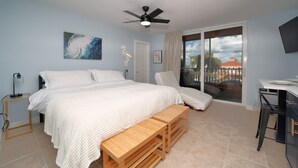 A bedroom with a bed, a desk, a chair, a bench, a ceiling fan, and a large window with a view of buildings. - Casa Playa #104 - Casa Flamingo - Pet Friendly (Fort Myers Beach)