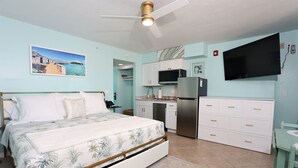 A bedroom with a bed, a ceiling fan, a TV, a kitchen area with cabinets and a refrigerator, and a bathroom with a shower. - Casa Playa #105 - Sea La Vie - Pet Friendly (Fort Myers Beach)