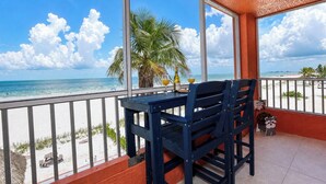 Un balcón con mesa y sillas, con vista a una playa con palmeras y el océano.