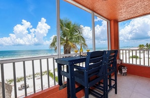 A balcony with a table and chairs, overlooking a beach with palm trees and the ocean.
