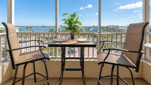 A balcony with two chairs and a table, a potted plant, and a view of the ocean and buildings. - Casa Playa #404 - Sandy Shores (Fort Myers Beach)