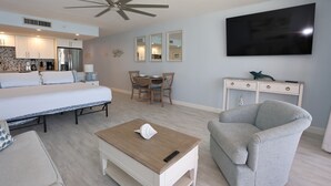 Ein modernes Hotelzimmer mit einem Bett, einem Holztischchen, einem grauen Sessel, einer Essecke mit Tisch und Stühlen, einer Küchenzeile und einem an der Wand montierten Fernseher.