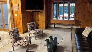 Una habitación con paneles de madera, un televisor, sillas estampadas, una mesa de centro de vidrio y un sofá de cuero.